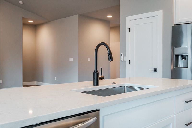 Large kitchen stainless steel sink and appliances, black faucet and hardware on doors and cabinets. Sink located on an island with off-white marble countertop with light gray veining, white cabinets, pantry with door, gray walls with white trim and can lighting