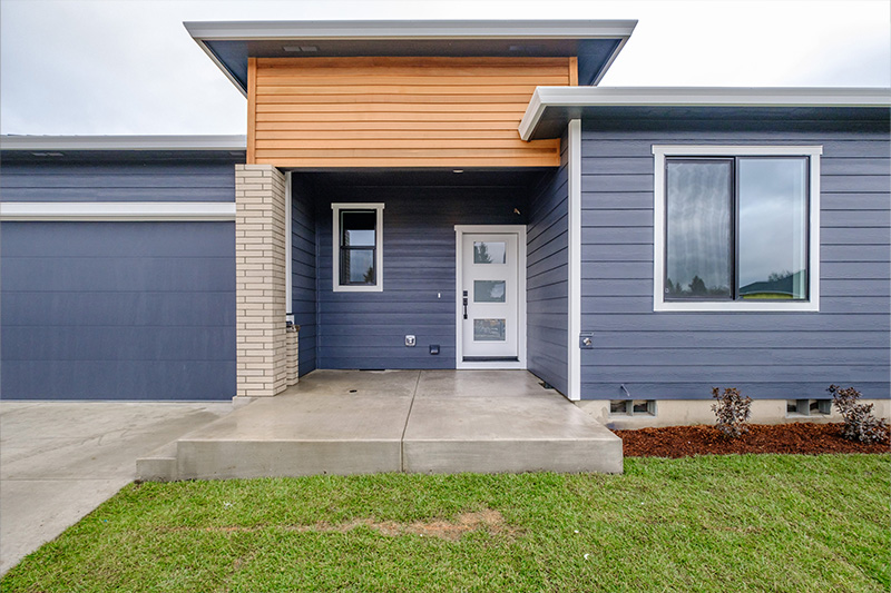 Front of a freshly built home with navy siding and garage door, brick and wood accents, concrete porch with small shrubs and brown mulch and grass