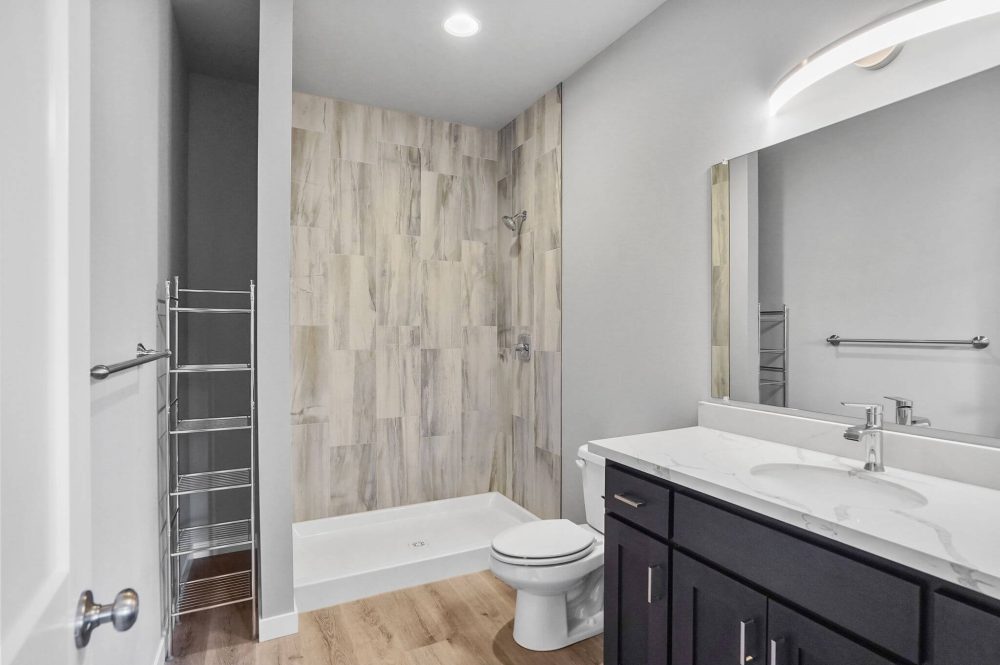 Large bathroom with standing shower lined with gray and cream colored tiles, stainless steel accents, fixtures and faucets, a toilet, towel rack, and gray cabinets and white marble with gray vein vanity