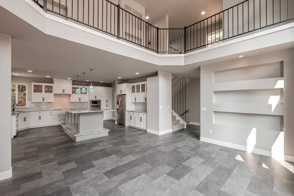large living room with small builtin fire place under builtin area for tv, built in white shelves on either side of tv and fireplace area, large kitchen with white cupboards and an island with gray tile floor, open upstairs walkway with black railing and stairs