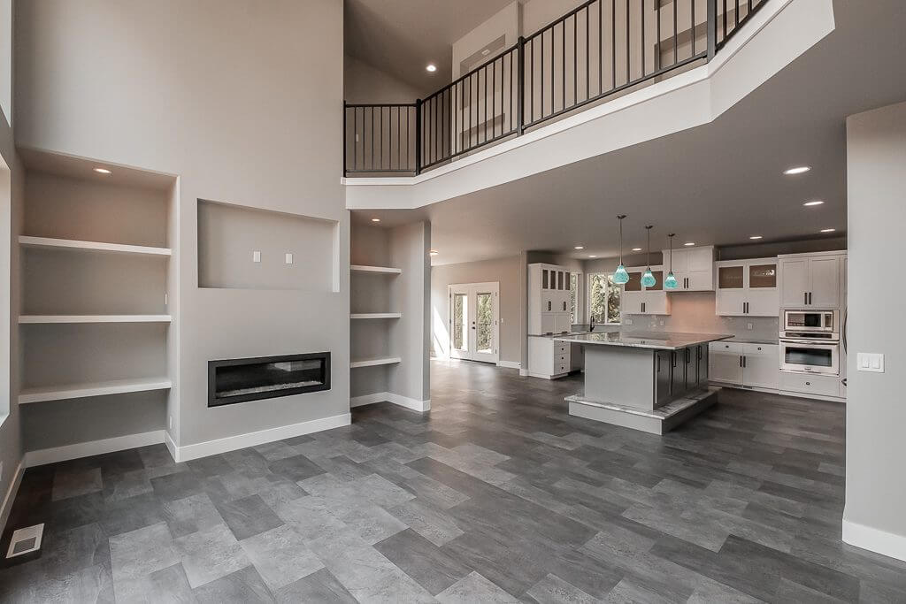 large living room with small builtin fire place under builtin area for tv, built in white shelves on either side of tv and fireplace area, large kitchen with white cupboards and an island with gray tile floor, open upstairs walkway with black railing