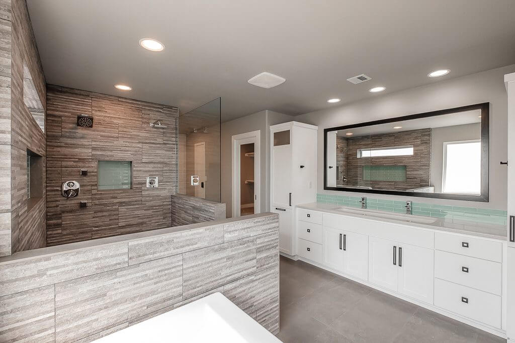 custom home large bathroom with a white bathtub, gray rock tiling throughout shower arear with dual stainless steel shower heads, turquoise brick backsplash accents in shower and along dual sinks with stainless steel faucets, white cupboards