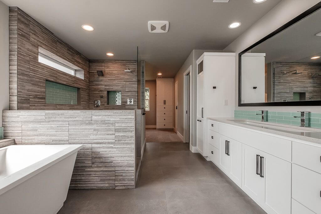 custom home large bathroom with a white bathtub, gray rock tiling throughout shower arear with dual stainless steel shower heads, turquoise brick backsplash accents in shower and along dual sinks with stainless steel faucets, white cupboards