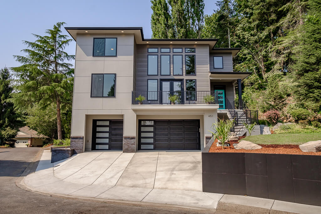 451 Mcnary exterior custom built multistory home with several large windows, stairs to turquoise front door, gray siding with black trim, a two-car and single car attached garages with trees and landscaping