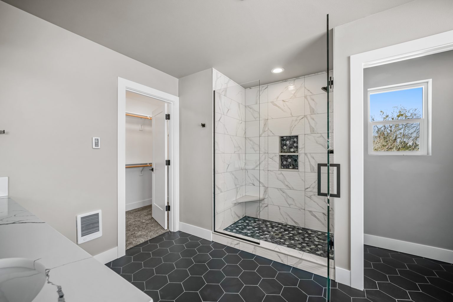 large bathroom with a standing shower that has a glass door and horizontal white marble tiles with gray veining, honeycomb dark floor tiles, bathroom sink and walk in closet