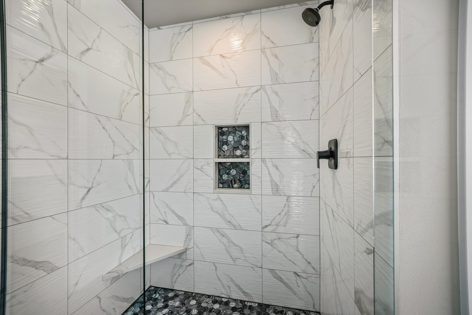 shower with black faucet and shower head, glass doors, and white tiles with gray veining that has two built-in shelves that have tile backsplash that matches the tile flooring