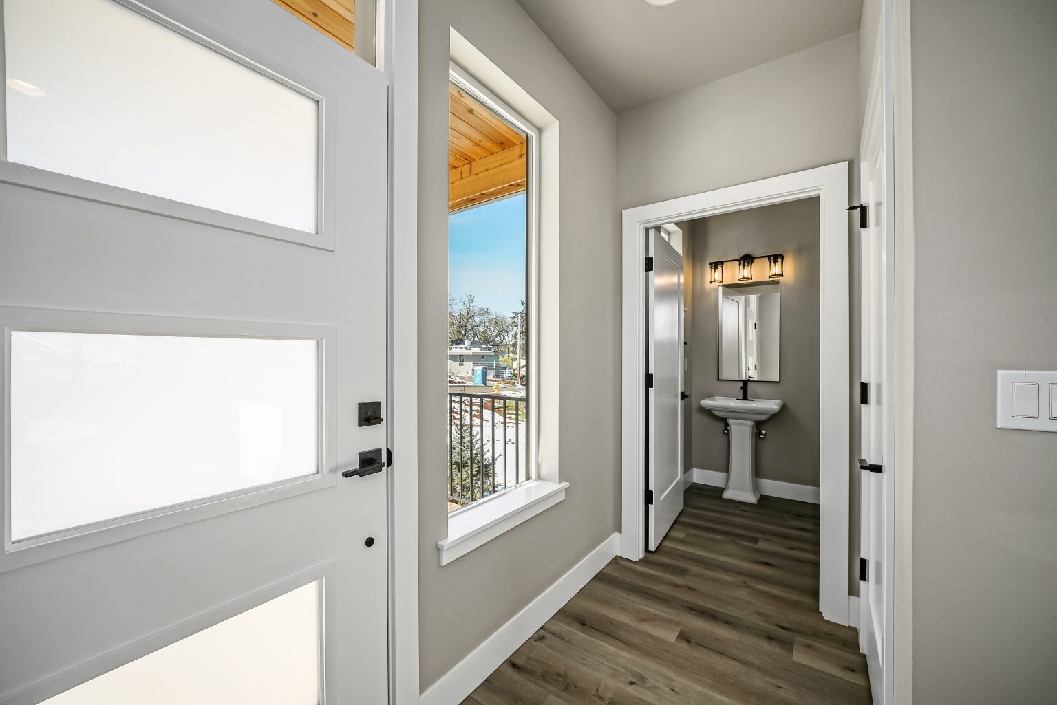 interior front door entry with large window, white trim, gray walls, hardwood flooring and opening to a half bathroom in the hallway