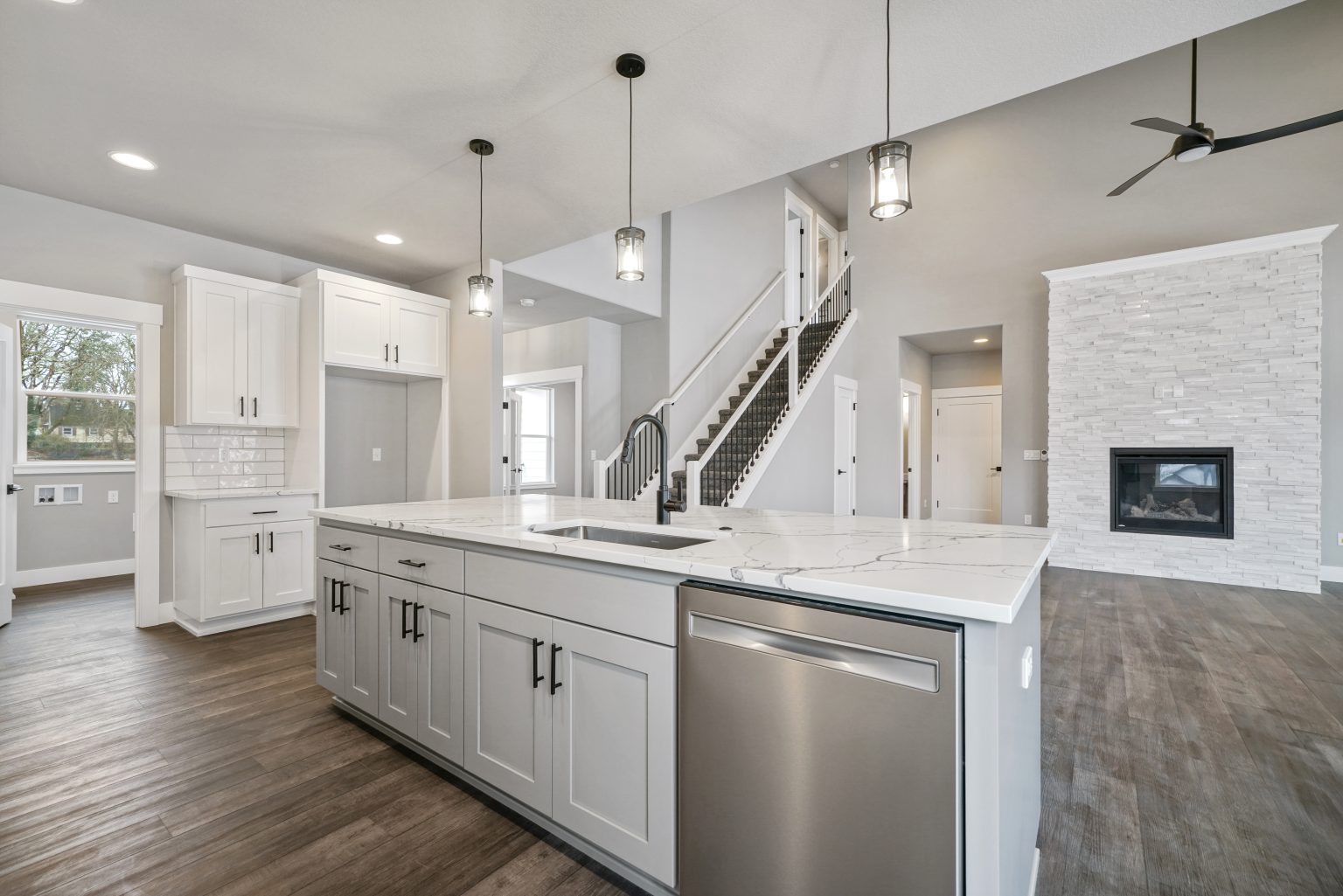 Beautiful, spacious open floor plan in the kitchen and living spaces, kitchen island, white cupboards stairs leading to top floor, and white exposed stone wall with built-in gas fireplace